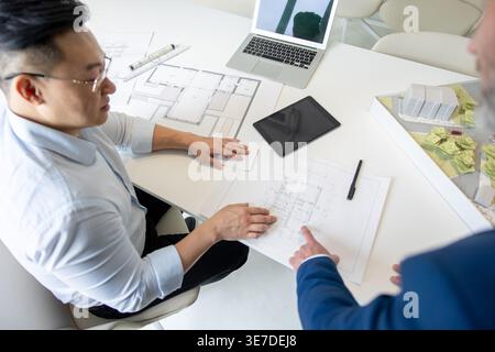 Asian male architect sitting and discussing blueprint with caucasian male colleague standing at table with architectural models, laptop, and tablet... Stock Photo