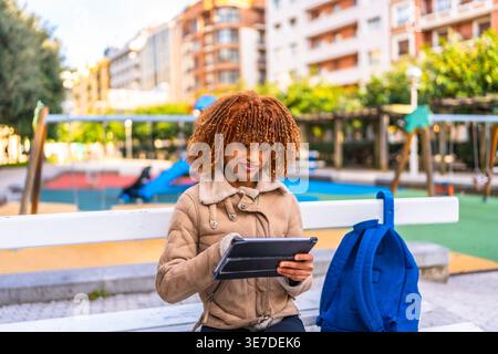 Mixed-race female teenager with blonde twists wearing brown jacket using tablet while sitting on bench with blue backpack in city park during daytime Stock Photo
