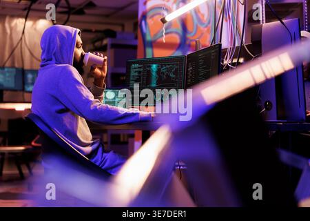 Hacker drinking coffee and doing illegal activities on computer late at night. Criminal holding beverage to go while cracking password and using malicious software in abandoned warehouse Stock Photo