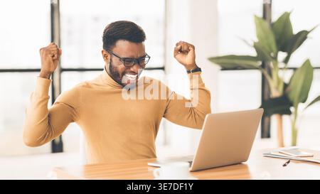 Cheerful Black man wearing eyeglasses raising hands sitting at table with laptop in office Stock Photo