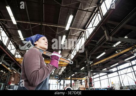 Black, Asian female worker looking away holding a wrench standing in factory Stock Photo