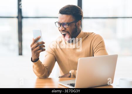 Angry Black man with eyeglasses having videocall using smartphone sitting at table with laptop in office Stock Photo