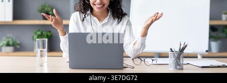 Black female teacher wearing headphones having online class on laptop sitting at table at home office Stock Photo