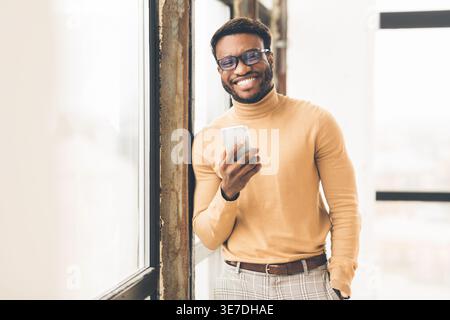 Smiling Black man with eyeglasses holding smartphone standing near window in office Stock Photo
