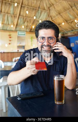 Smiling Hispanic middle-aged man with eyeglasses talking on smartphone and handing credit card sitting at table in restaurant Stock Photo