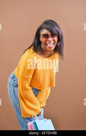 Cheerful Black woman in yellow ribbed long-sleeve crop top and sunglasses holding shopping bags and laughing while standing against brown backgroun... Stock Photo