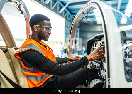 African American male aircraft engineer wearing reflective vest sitting inside private jet checking cockpit control system in hangar Stock Photo