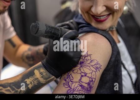 Cropped image of Asian male tattoo artist in latex gloves working with tattoo machine putting ink on arm of middle-aged caucasian female client in ... Stock Photo