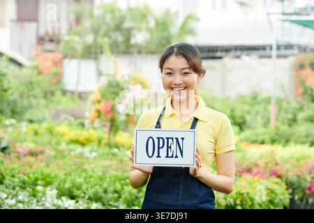 Smiling Asian female worker holding open signage standing in plant market Stock Photo