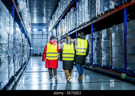 Diverse group of logistics team in safety vests inspecting pallets of products wrapped in plastic on high racks while walking down the aisle in col... Stock Photo