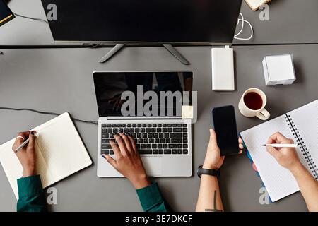 Black female programmer taking notes and using laptop near Asian male colleague holding pen and smartphone with codes on screen sitting together at... Stock Photo