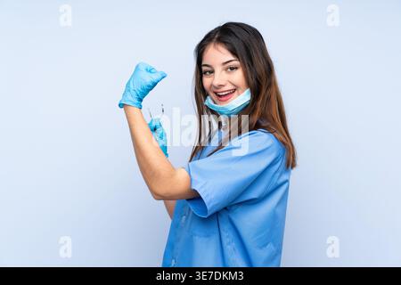 Hispanic female dentist holding dental mirror and sickle probe and showing arm muscle while standing against light blue background Stock Photo