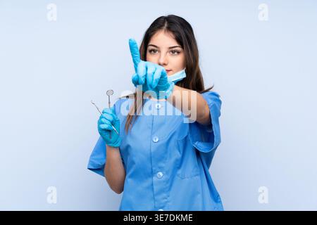 Hispanic female dentist holding dental mirror and sickle probe pointing finger away while standing against light blue background Stock Photo