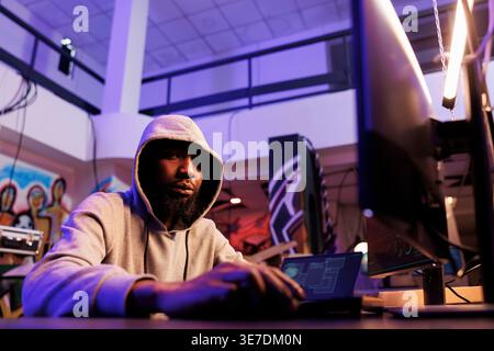 Hacker coding internet virus malicious software on computer portrait. African american man in hood doing ddos attack illegal activity and breaking law while looking at camera Stock Photo