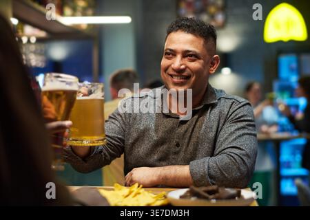Smiling mixed race man in casualwear having toast of beer with friend sitting at a table in bar Stock Photo