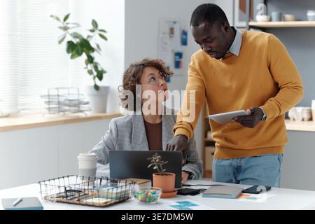 Black man pointing laptop screen giving instructions to caucasian female colleague sitting at table with laptop in office Stock Photo