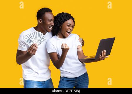 Cheerful Black man holding bunch of money looking at laptop of his girlfriend while standing against yellow background in studio Stock Photo
