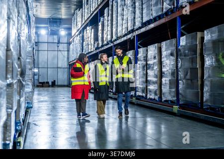 Diverse group of logistics team in safety vests discussing inventory management while walking with barcode scanner and clipboard in cold storage wa... Stock Photo