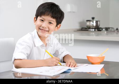 Smiling Chinese boy doing homework sitting at table in kitchen at home Stock Photo