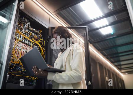 Black female IT engineer wearing eyeglasses using laptop while standing near server cabinet in data center Stock Photo
