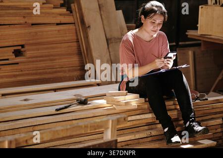 Asian female carpenter with clipboard on lap using smartphone while sitting on wooden planks in workshop Stock Photo