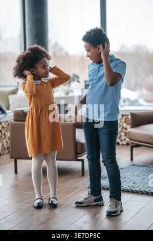 Smiling Black siblings looking at each other while touching hair standing in living room at home during daytime Stock Photo