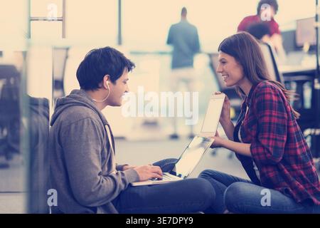 Smiling Mixed race man with earphones using laptop with caucasian female coworker showing tablet sitting on floor in office Stock Photo