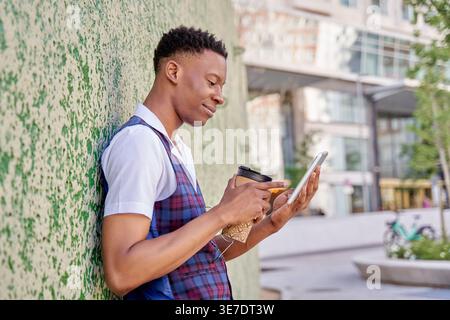 Black man holding takeaway coffee and using tablet while standing and leaning against concrete wall in city during daytime Stock Photo