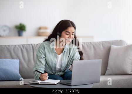 Indian female in headset writing down notes having online class on laptop sitting on sofa at home Stock Photo