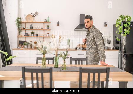 Arab military man with sad facial expression wearing camouflage uniform carrying backpack over shoulder standing at wooden kitchen table at home Stock Photo