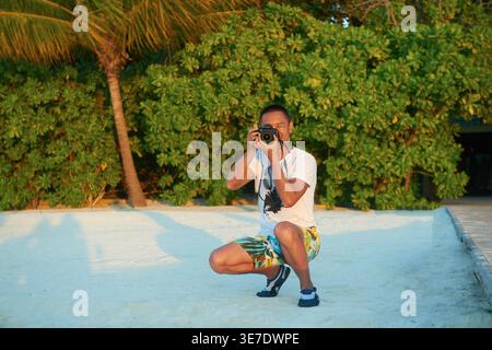 Asian man taking pictures using photo camera while crouching on beach against green tropical plants and tree on sunny day Stock Photo