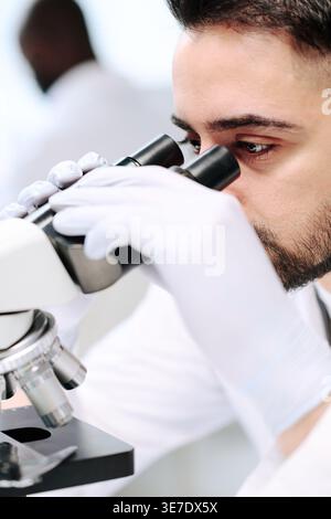 Middle Eastern man wearing white lab coat and gloves investigating bacteria using microscope in laboratory Stock Photo