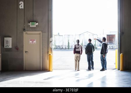Asian American male pointing away while talking to co-workers standing in warehouse Stock Photo