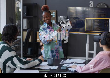 Black female programmer with high bun standing and presenting new VR glasses to her colleagues during team meeting in office Stock Photo