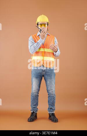 Serious Asian male contractor in orange vest, goggles, and hardhat standing while talking on walkie-talkie against orange background in studio Stock Photo