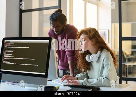 Indian male programmer standing near caucasian female colleague working on computer sitting at table in office Stock Photo