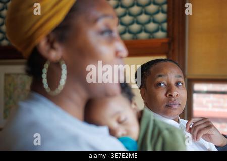 Black non-binary person standing with wife carrying their baby in the living room at home Stock Photo