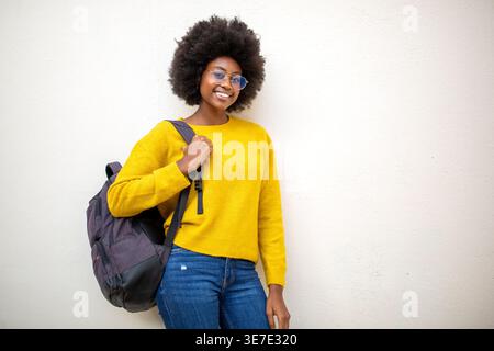 Smiling Black woman with afro wearing eyeglasses holding backpack strap while standing against white background Stock Photo
