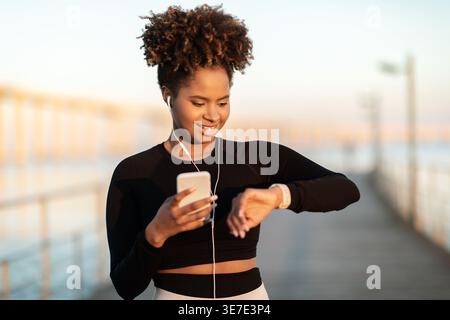 Black woman with afro wearing workout shirt and earphones holding smartphone and checking time on her watch while standing on boardwalk at dawn Stock Photo