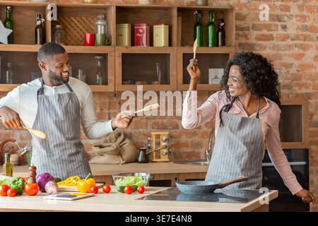 Cheerful Black couple wearing aprons having fun playing with wooden utensils while standing at kitchen counter with vegetables at home Stock Photo
