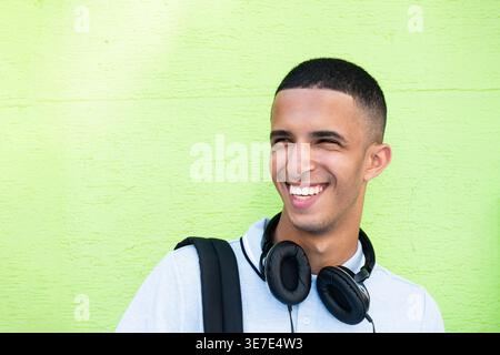 Cheerful Arab male with headphones on neck standing against green wall during daytime Stock Photo