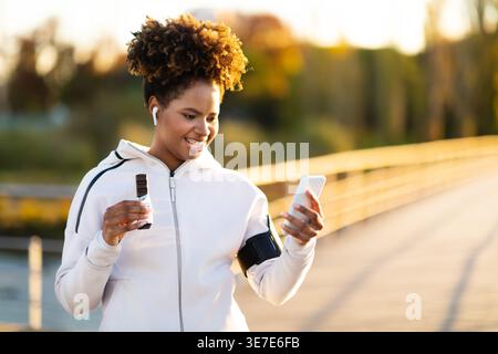 Black woman with afro and dental braces wearing white jacket and wireless earphones using smLifestylephone and holding protein bar while standing outdoor... Stock Photo