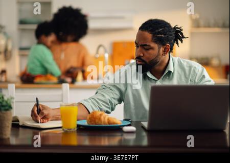 Serious Black father sitting at table with laptop writing notes working remotely near wife and son playing at kitchen counter at home Stock Photo
