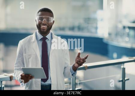 Black male medical scientist in labcoat and protective glasses gesturing hand holding tablet standing in laboratory Stock Photo