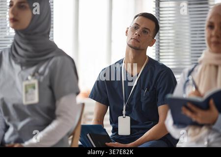 Mixed-race male doctor in scrubs wearing eyeglasses holding notebook and folder looking away while sitting on chair near female colleagues during m... Stock Photo