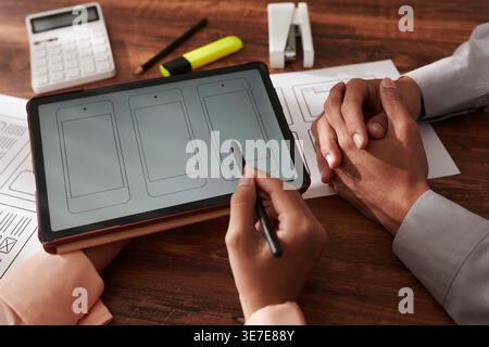 Asian male designer and woman colleague discussing application mockup on tablet while sitting at table with documents in office Stock Photo