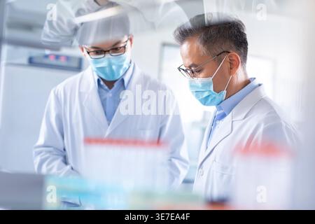 Chinese male researchers wearing lab gowns and face masks standing with clipboard working together in laboratory Stock Photo