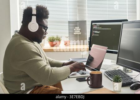 Serious Black male programmer wearing wireless headphones using laptop writing computer codes to program while sitting at table with coffee in office Stock Photo
