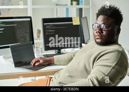 Serious Black male programmer wearing eyeglasses sitting at table with laptop and computers in office Stock Photo