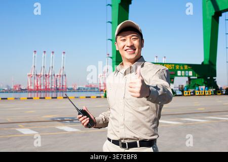 Smiling Chinese male shipping industry worker wearing white hardhat holding hand-held radio and showing thumbs up while standing at a shipping port Stock Photo